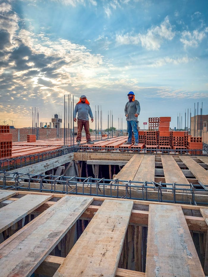 Two construction workers stand on a building site under a cloud-filled sky. Sunlight illuminates the scene.