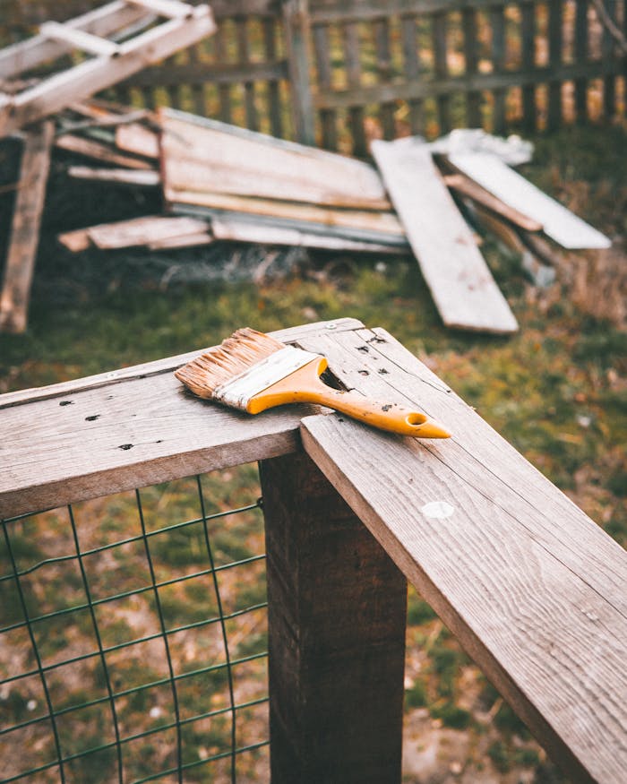 A paintbrush rests on a rustic wooden fence in a yard, suggestive of outdoor home maintenance or DIY project.